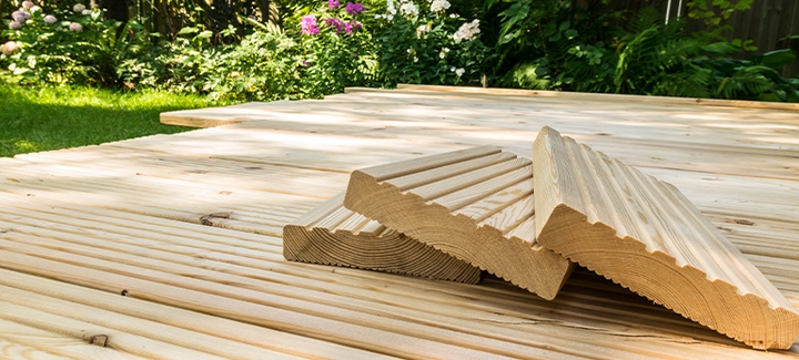 Terrassenbau mit hellen Lärchenholzdielen, gestapelte Riffelbretter im Vordergrund, umgeben von grünem Garten.