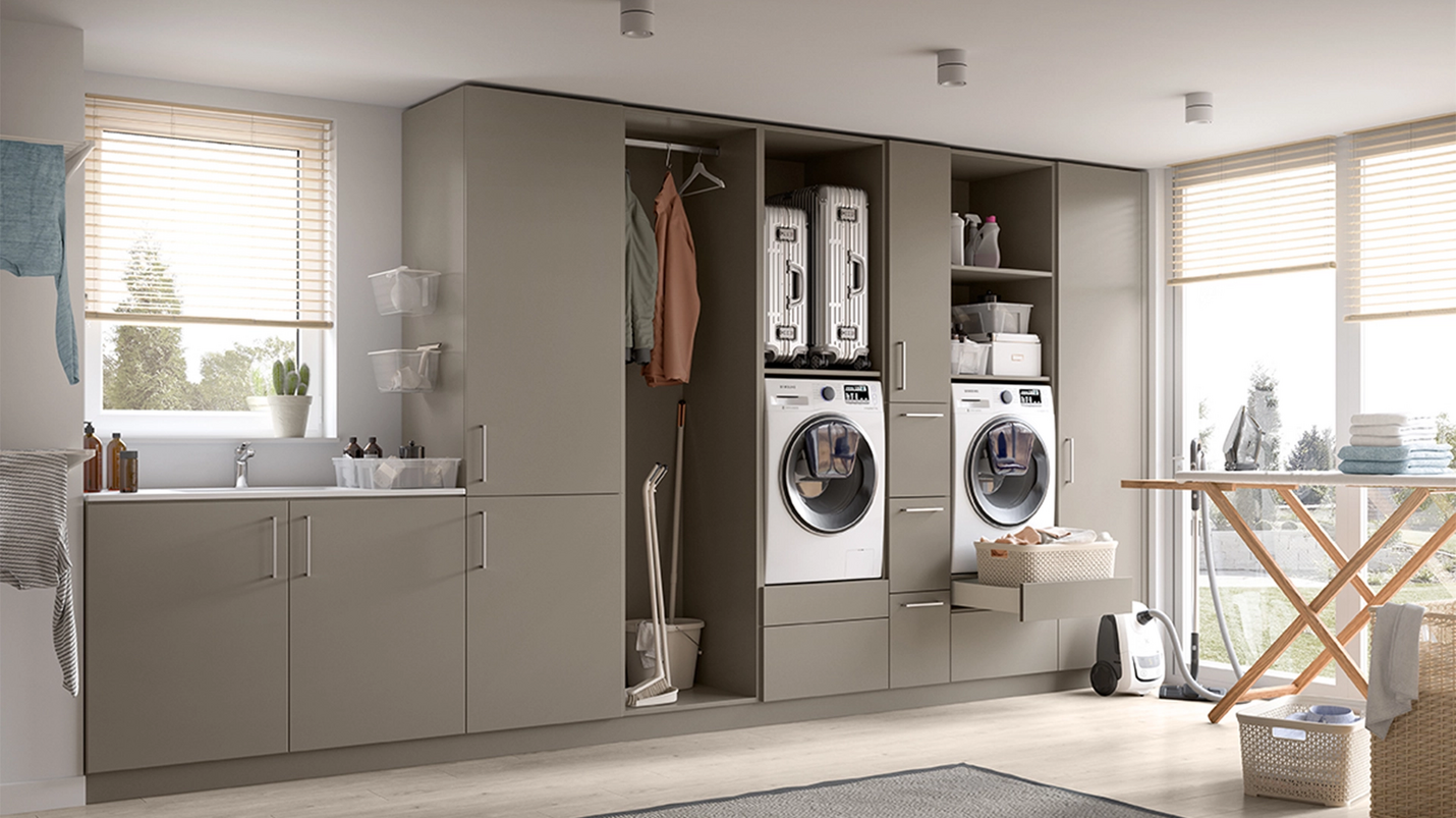 Moderner Hauswirtschaftsraum mit grauen Einbauschränken, Waschmaschine und Trockner, viel Stauraum und Bügelbrett am Fenster.