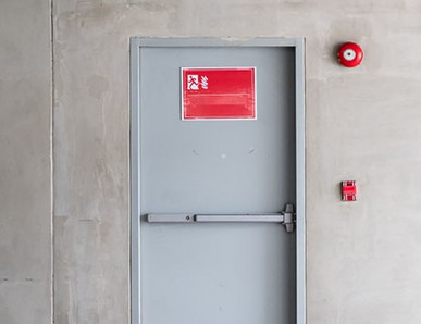 Graue Brandschutztür mit Fluchtwegsymbol und roter Alarmglocke.